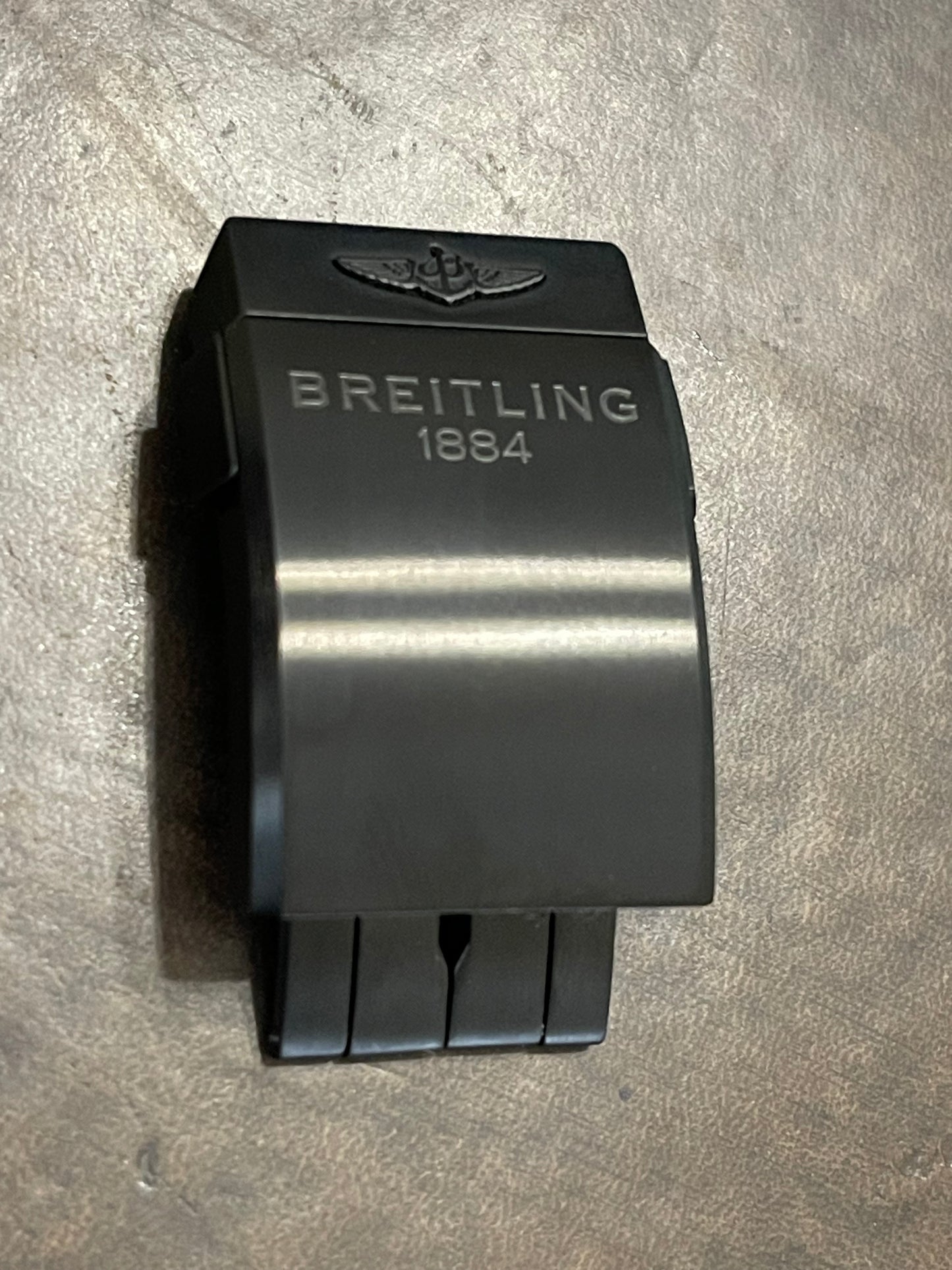 Close-up of a black Breitling watch clasp engraved with "Breitling" and "1884," resting on a textured surface.