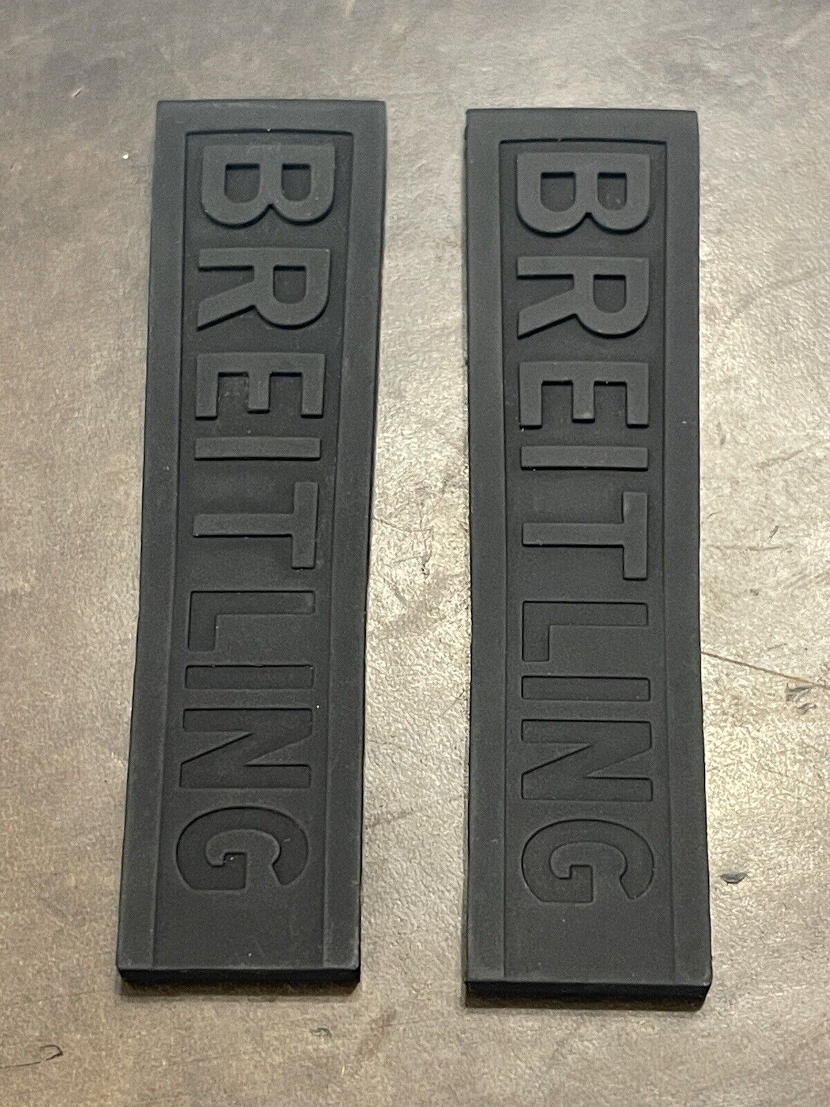 Black rubber watch strap parts with 'BREITLING' text side by side on rough background